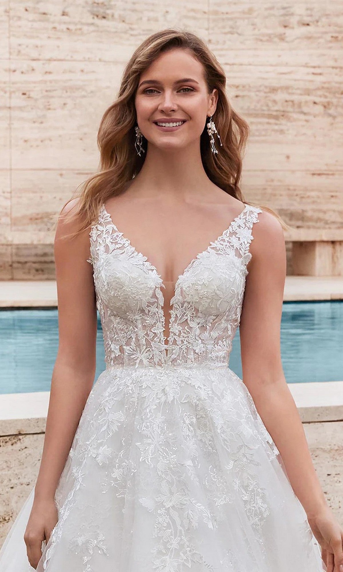 A woman in the Alyce 7039 long white dress, featuring a sleeveless lace ball gown design with a deep V neckline, smiles at the camera while standing by a pool and stone wall.